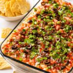 7 Layer Tex Mex Dip in a glass bowl showing distinct layers of beans, cheese, sour cream, guacamole, and salsa.