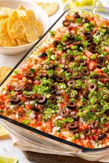 7 Layer Tex Mex Dip in a glass bowl showing distinct layers of beans, cheese, sour cream, guacamole, and salsa.