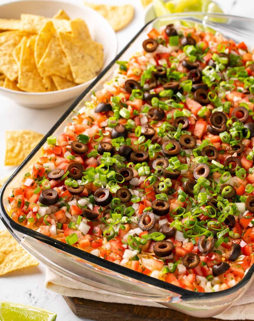 7 Layer Tex Mex Dip in a glass bowl showing distinct layers of beans, cheese, sour cream, guacamole, and salsa.