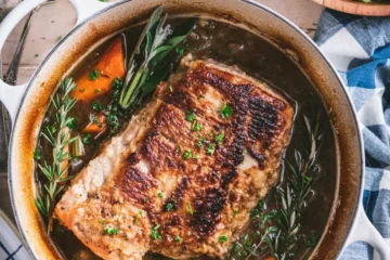 A tender hunk of beer-braised pork shoulder in a Dutch oven, surrounded by carrots, onions, and herbs.