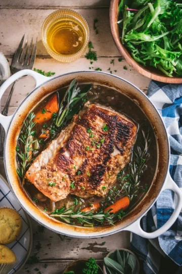 A tender hunk of beer-braised pork shoulder in a Dutch oven, surrounded by carrots, onions, and herbs.